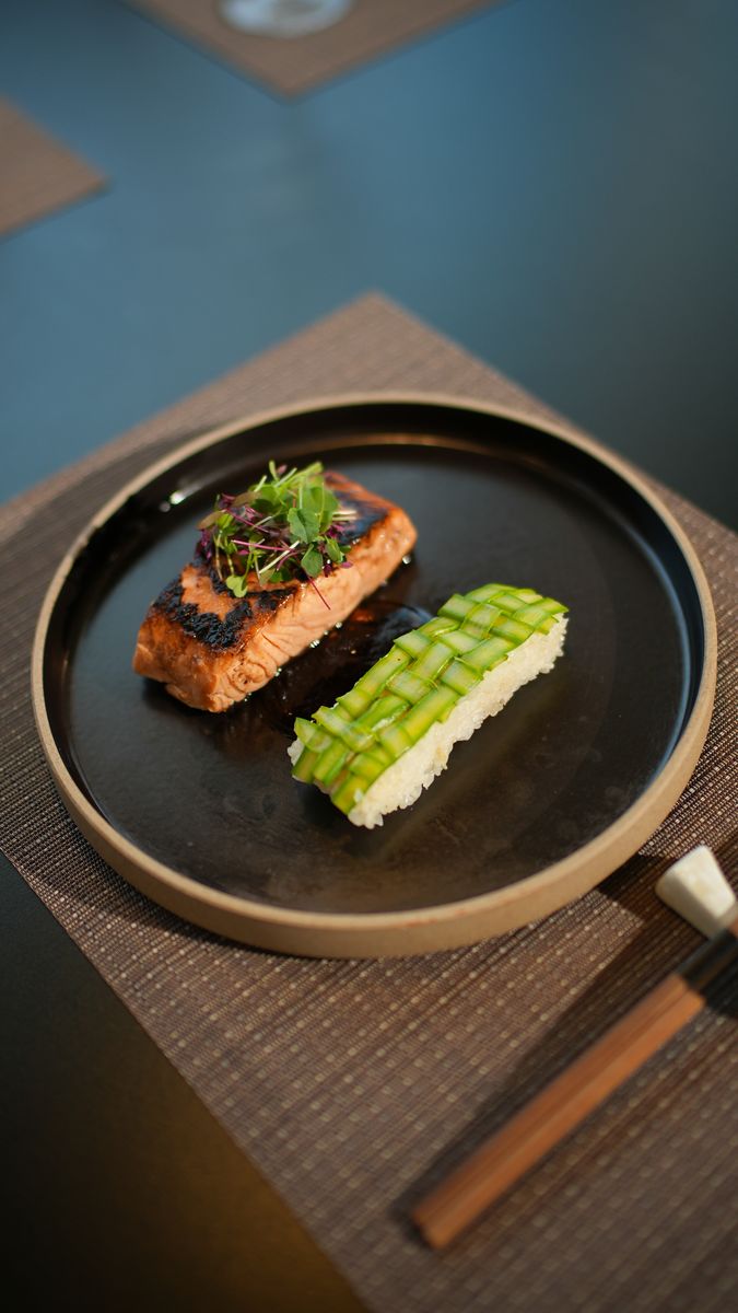 Seared salmon beside a green roll on a black plate
